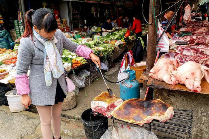 在一个猪肉摊点前，这个卖猪肉的小大姐用液化气做火源烧烤大块的猪肉的皮。