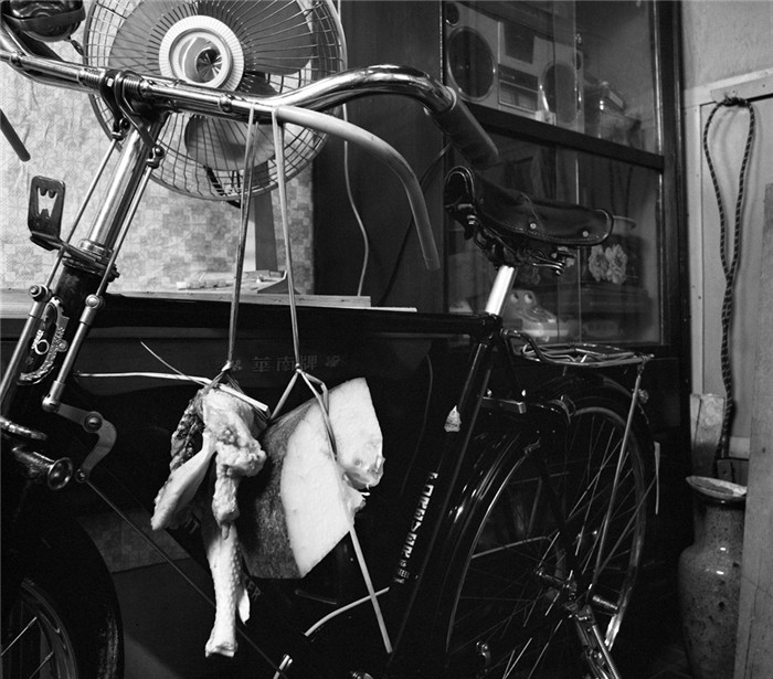 图为1982年，广东广州一个居民家门口，自行车上悬挂着买回来的猪肉，自行车后面的桌上摆放着电风扇，柜子里放着录音机——这是典型的80年代家电画面。 